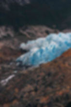 A blue glacier sits on rocky terrain with dark mountains in the background.