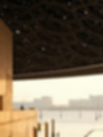 A person stands on a balcony by water at the Abu Dhabi Louvre, with a unique patterned ceiling above and city buildings in the background.