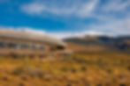 A modern, curved wooden building sits on a grassy plain, with mountains and snowy Patagonian peaks in the background under a blue sky.