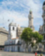 Historical buildings in Buenos Aires with a clock tower and white façade, surrounded by trees and pedestrians, under a partly cloudy sky.