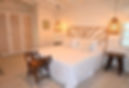 Cozy bedroom with a white bed, wicker headboard, and pendant lights. Wooden stools and a small bench add rustic charm. Neutral tones create a calm ambiance.
