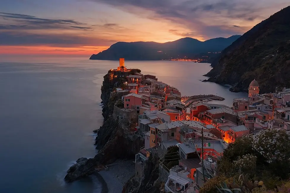 A scenic view of Vernazza at sunset, featuring colourful houses on a cliff by the sea, with a historic tower and glowing lights.