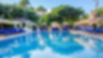 A serene poolside setting with clear blue water, surrounded by vibrant green palms. Blue lounge chairs and beige umbrellas create a relaxed, inviting atmosphere.