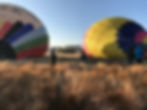Two colourful hot air balloons are inflated on a grassy field, with people preparing nearby in the early morning light in Morocco.