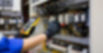 A person wearing black gloves uses a multimeter to measure the voltage of a large electrical panel on a wall.