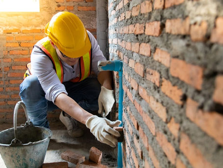 A worker carefully working on a brick wall, inspecting and handling the bricks while wearing a hard hat and gloves.