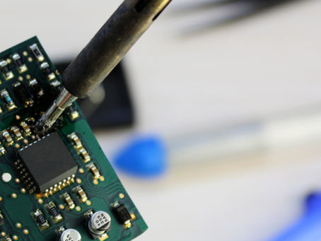 A soldering iron touching a green circuit board with black chips, silver capacitors, and blurred tools behind it.