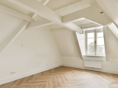 In a large empty room, there are sloped ceilings and large windows, and the floor has a large herringbone pattern.