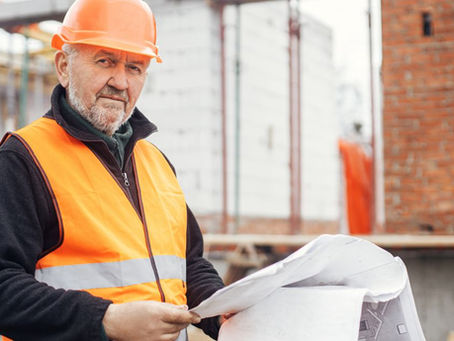 A worker wearing an orange high-visibility vest and helmet supervises a project and looks at blueprints.