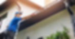 A worker on a ladder wearing gloves is cleaning out the black gutters on a white home.
