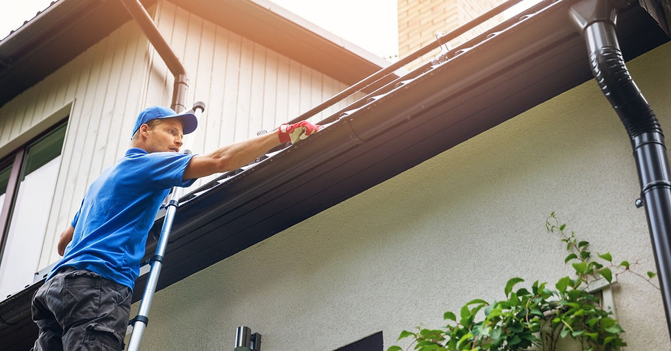 A worker on a ladder wearing gloves is cleaning out the black gutters on a white home.