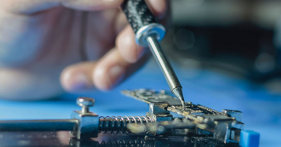 A hand holding a soldering iron over a small circuit board secured in a clamp on a blue work surface.