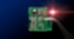A miniature controller circuit board is held up with tweezers against a dark background. A red light is shining on the board.
