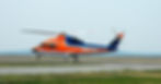 A blue and orange air ambulance helicopter, surrounded by grass and parked on an empty aircraft runway.