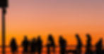 The silhouette of multiple people against a railing during a sunset flooding the sky with orange and yellow colors.