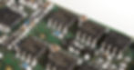 Close-up of a green circuit board with black integrated circuits, silver solder pads, and small blue and beige components.