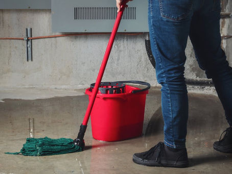 Someone stands in a puddle of water in a basement. They use a mop to soak up water with a red bucket nearby.
