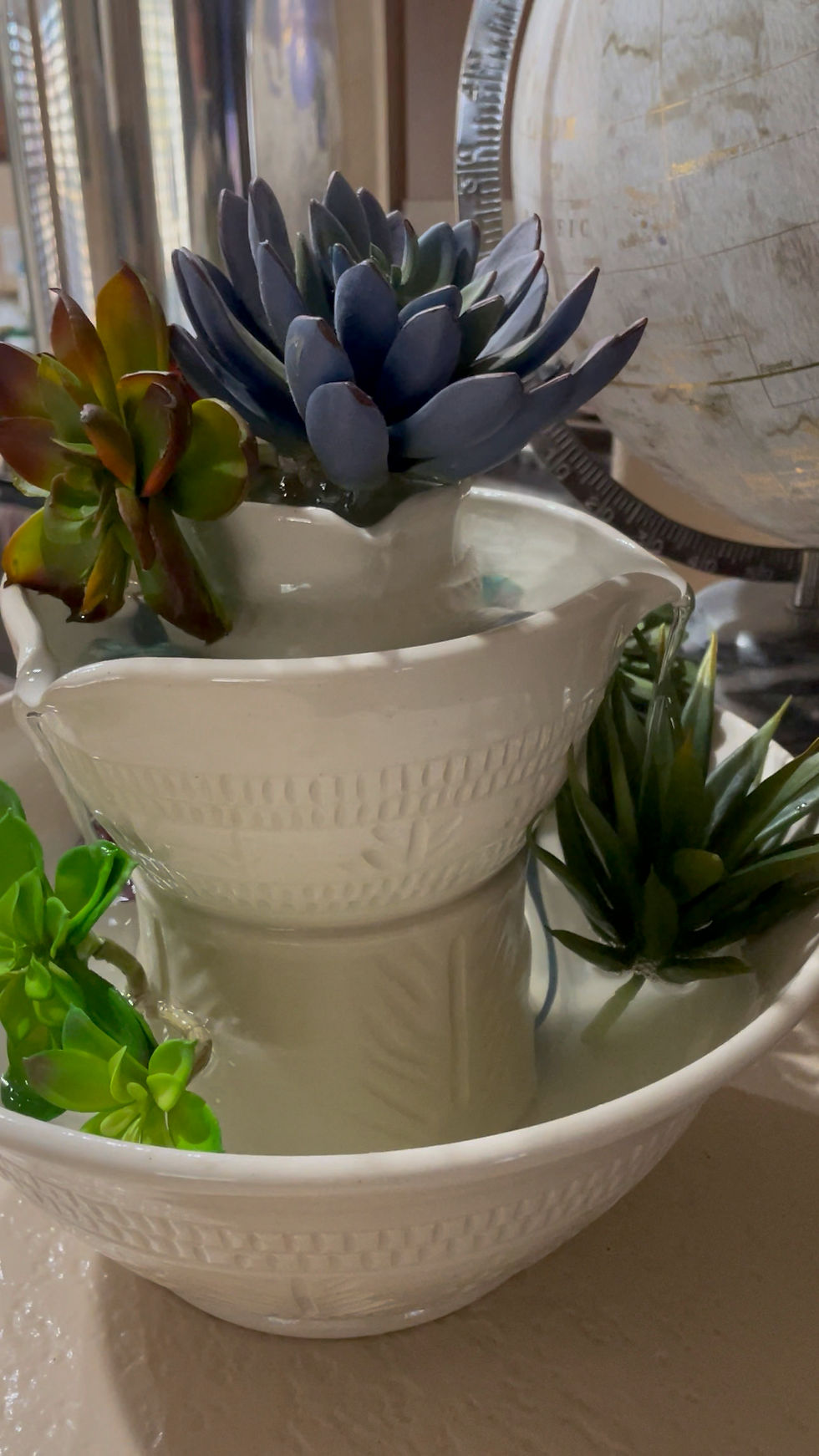 White ceramic tabletop fountain with colorful succulents indoors, beside a globe, creating a calm decorative display.
