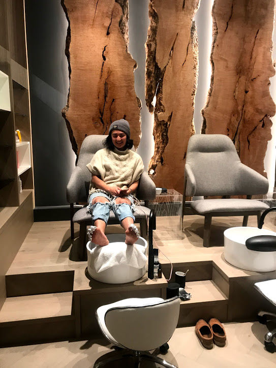 A girl, sitting in comfty pedicure station, with gorgeous wood carvings behind her