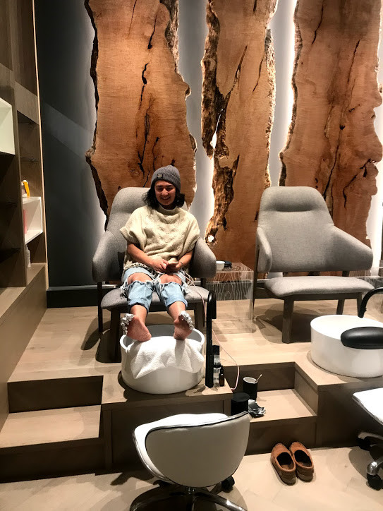 A girl, sitting in comfty pedicure station, with gorgeous wood carvings behind her