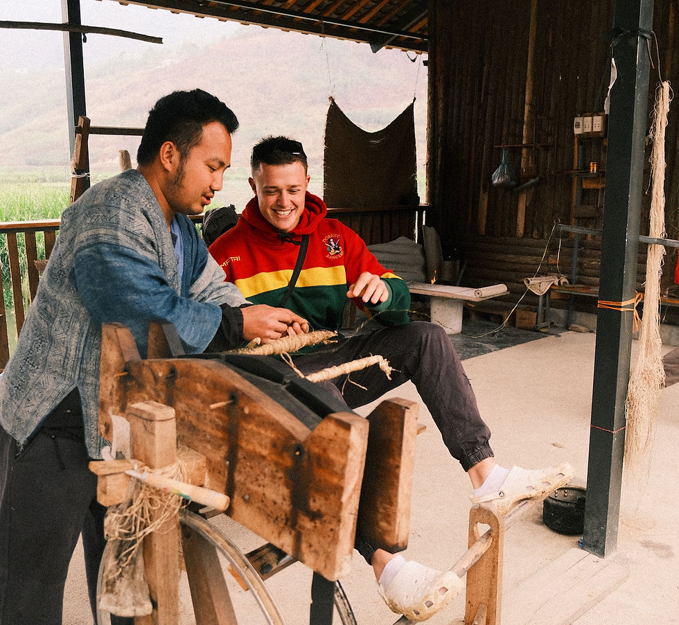 Two men are weaving yarn, one in a colorful hoodie. They sit in a rustic setting, smiling, amidst wooden tools and natural fibers.