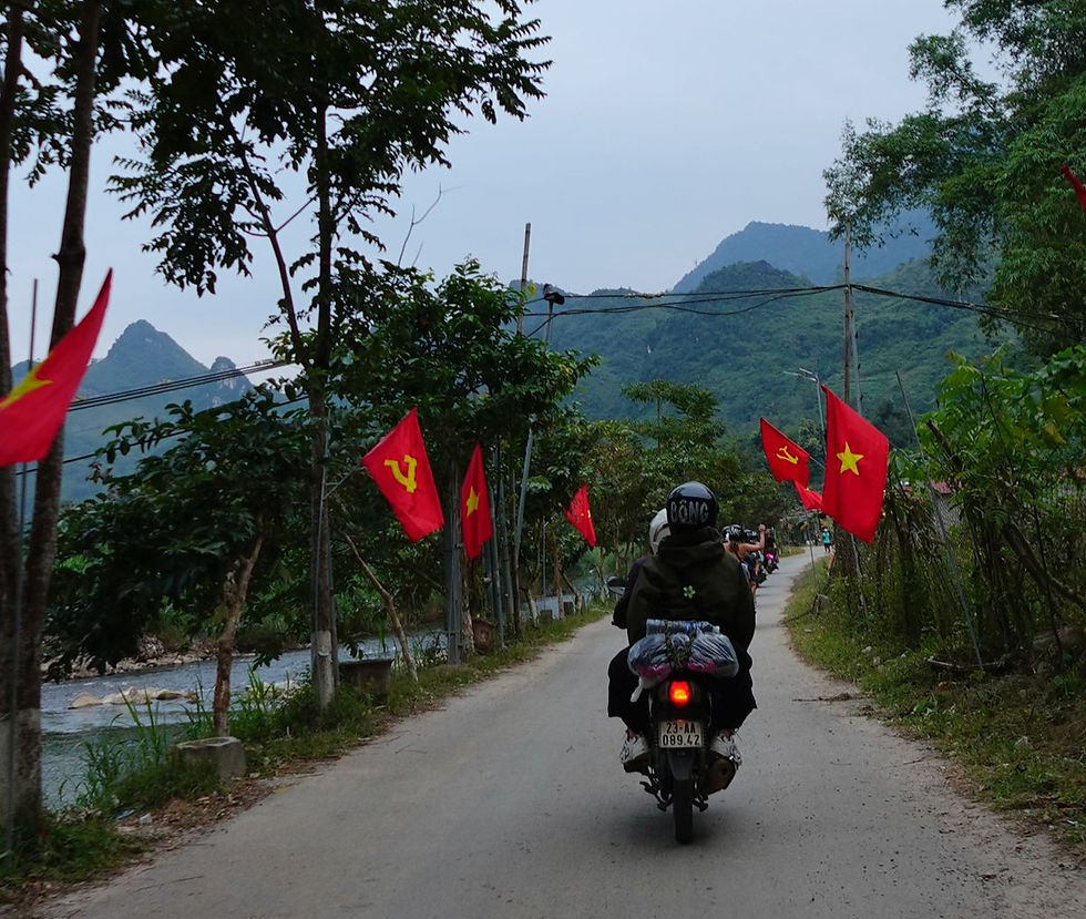 Motorcyclist rides on a narrow road lined with red flags featuring yellow stars and hammer-and-sickle symbols, set against lush mountains.