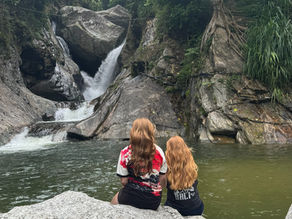 Two people with reddish hair sit on rocks by a waterfall surrounded by lush greenery. They're gazing at the water, creating a serene mood.