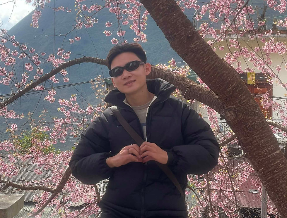 A person in a black jacket and sunglasses makes a heart gesture under pink cherry blossoms, with a mountain and building in the background.
