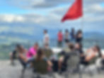 People relax on patio chairs overlooking scenic mountains, with a red flag nearby. Some enjoy drinks, others admire the view under cloudy skies.