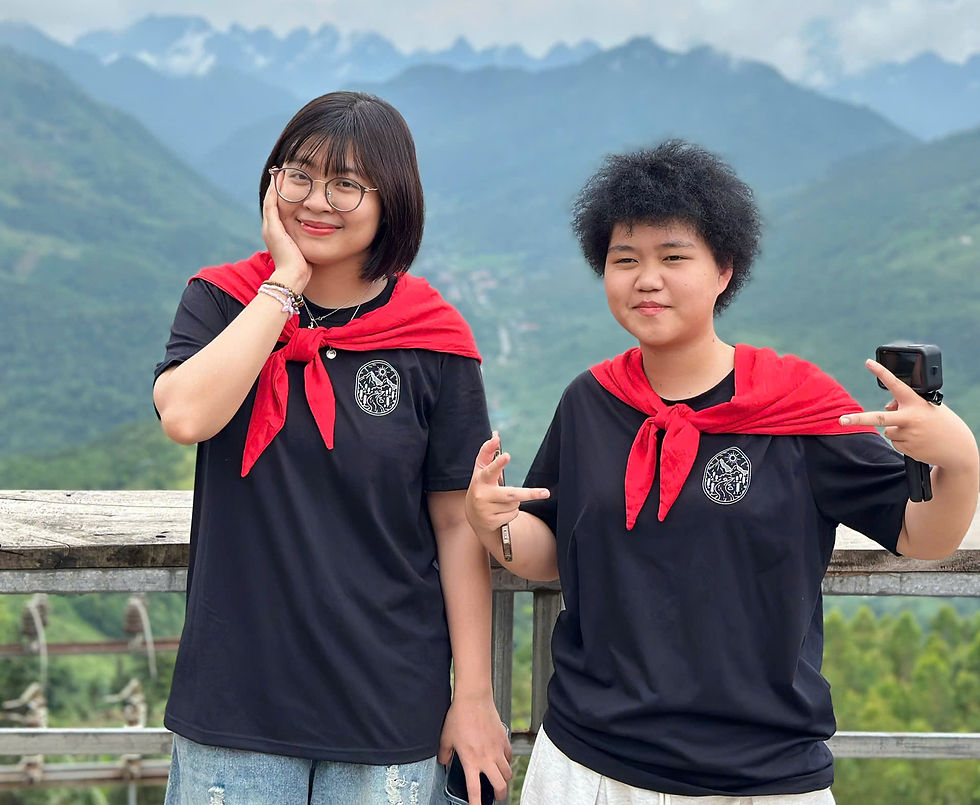 Two people in black shirts with red scarves pose on a mountain balcony. One smiles, the other makes peace signs. Cloudy sky and green hills.