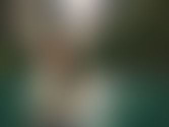 Smiling woman in a gray jacket stands near a turquoise river, flanked by tall cliffs. Boats are visible in the background. The mood is joyful.