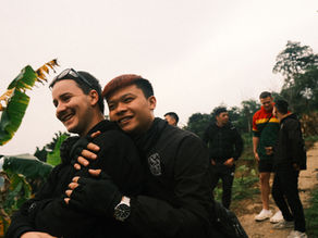 Two people smiling and hugging on a dirt path with banana trees. Three others chat in the background, wearing casual clothing.