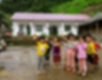 Group of children smiling and waving outside a white house with a red roof. Green hills in the background. Cheerful, vibrant scene.