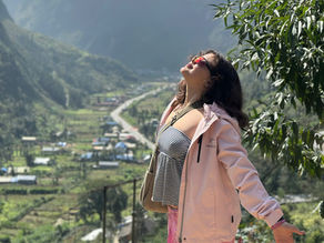 Woman in a pink jacket and striped top joyfully stretches arms out in a mountainous valley. Sunlight and greenery create a vibrant scene.