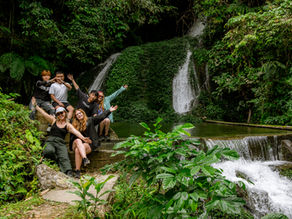 Group of six people posing with arms raised, smiling by lush green waterfall. Casual attire, vibrant foliage, joyful mood.
