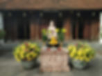 Stone statue surrounded by yellow lilies and fruit offerings on a carved pedestal. Wooden temple with intricate doors in the background.