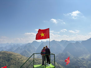 Couple stands on a scenic overlook with a Vietnamese flag, surrounded by lush, mountainous landscape under a clear blue sky.