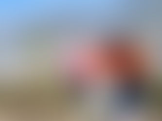 Three women in warm clothes sit smiling on a mountain ledge, with scenic hills in the background under a clear blue sky.