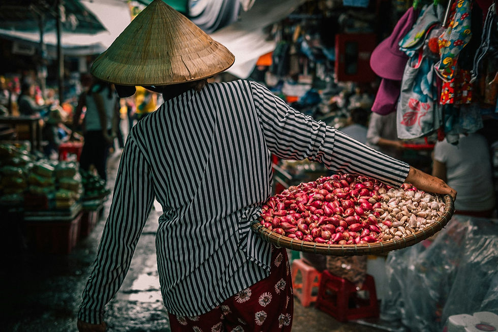 A person in a striped shirt and conical hat carries a basket of red onions in a bustling, colorful market setting.