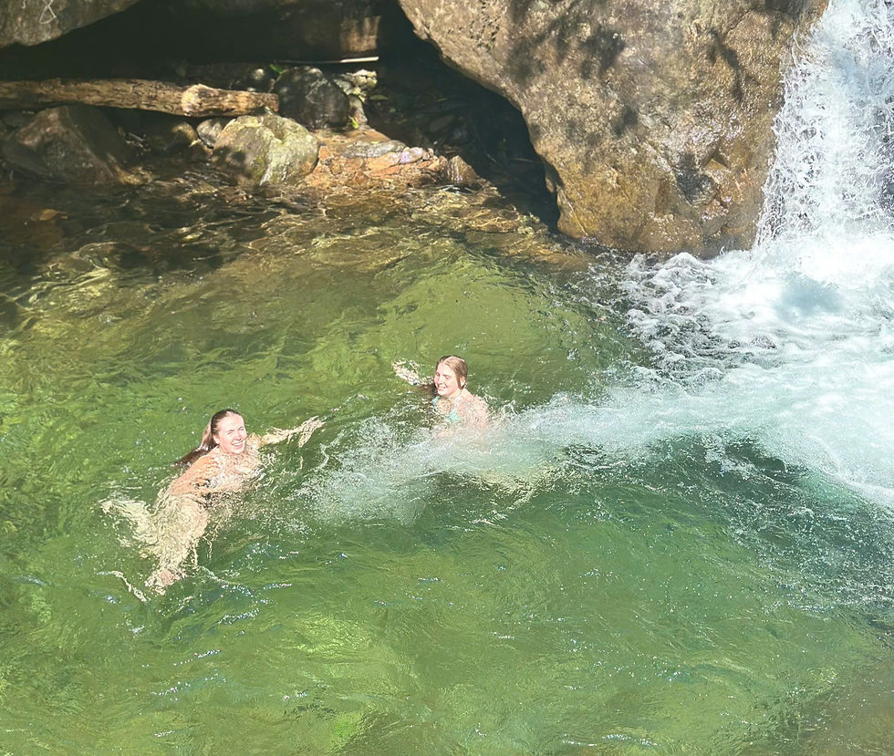 Two people joyfully swim in clear green water near rocks and a waterfall under bright sunlight, creating a lively and refreshing scene.