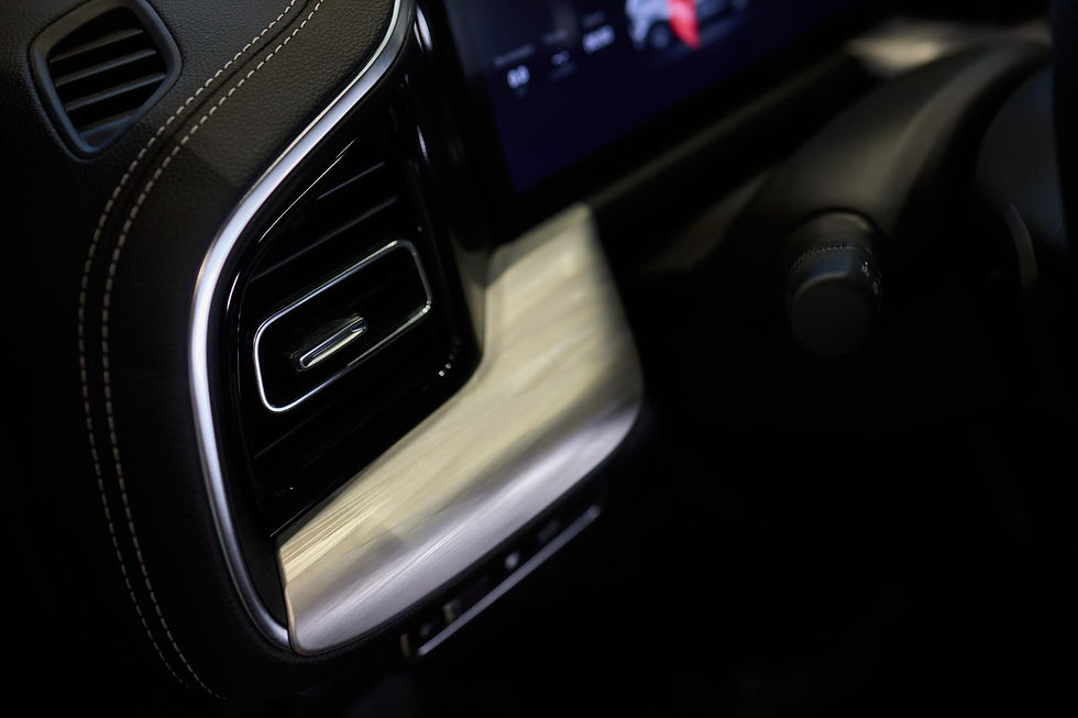 A close-up of a car's interior shows a sleek air vent with a metallic trim, positioned next to a light-colored wooden dashboard panel and a black rotary knob with textured edges.