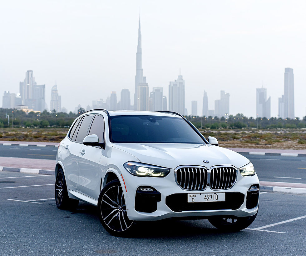 A white BMW SUV with sleek design features is parked on a road with the Dubai skyline, including the Burj Khalifa, visible in the background under a clear sky.
