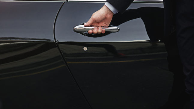 A person in a suit opens the door of a sleek black car, with their hand on the shiny silver handle.