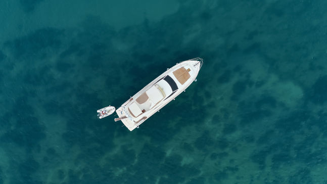 Aerial view of a sleek white yacht with beige accents floating on clear blue-green water, accompanied by a small dinghy tied to its side.