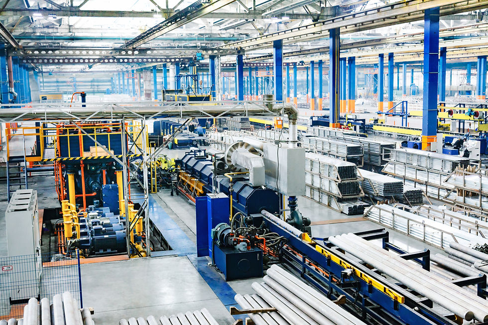 A high-angle view of a large manufacturing and industrial facility floor featuring heavy assembly machinery, overhead structural beams, and neatly organized stacks of raw metal pipes and parts, illustrating the complex supply chain and parts procurement operations optimized by Quidi's sourcing solutions.