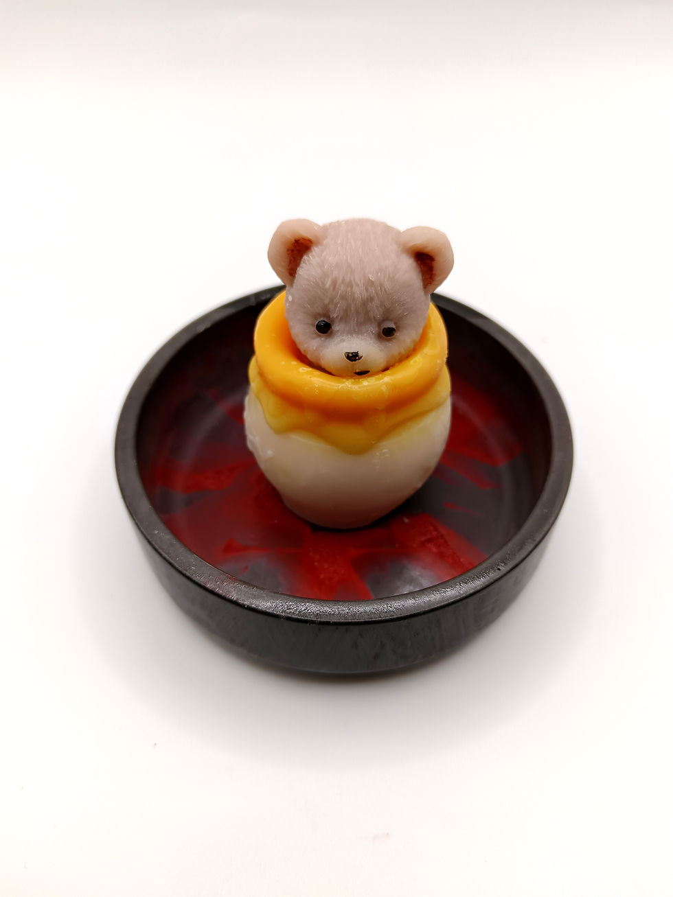 Fondant Honey Boom parfum cire et miel en forme de petit ours beige posé dans une coupelle ronde sombre au fond rouge.