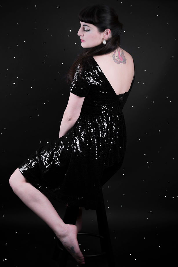 Portrait en pied, photographie couleur d'une performeuse, pin-up en robe de soirée noire brillante sur fond étoilé.