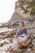 Petra Beumer, founder of Mindful Eating Institute in Santa Barbara, in white pants and purple shawl sits on rocks at a rocky beach beneath a cliff, gazing calmly out to sea.
