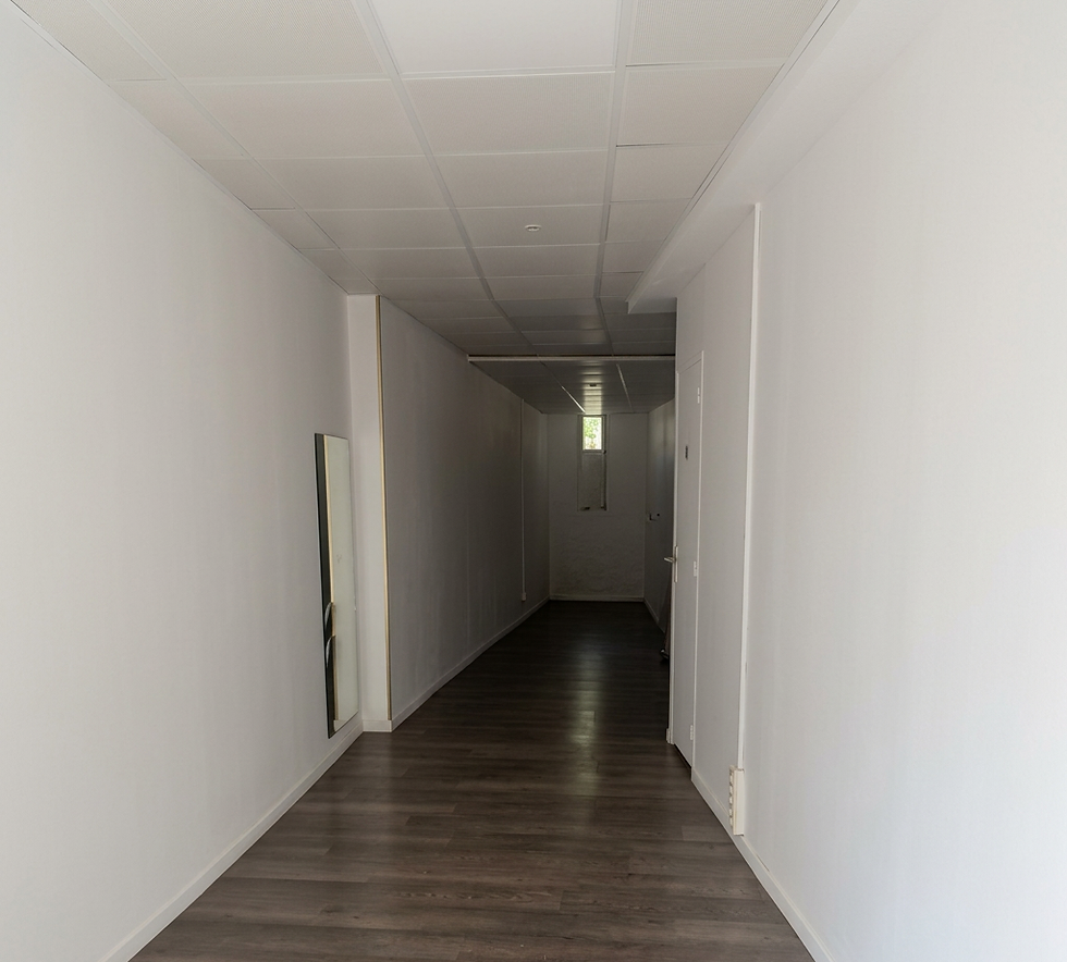 Interior photography of a long, narrow hallway with white walls and a dark wooden floor. The perspective leads towards a bright light at the far end, suggesting a path or transition.