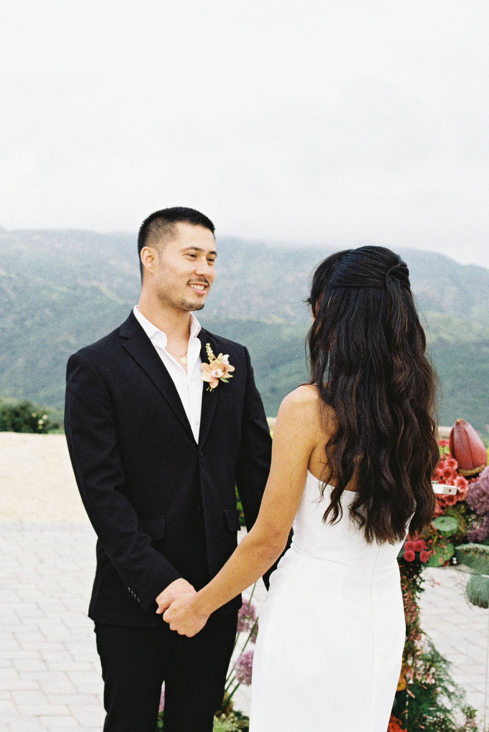 Bride and groom holding hands during an outdoor ceremony with mountain views and ocean veiws and colorful floral arrangements